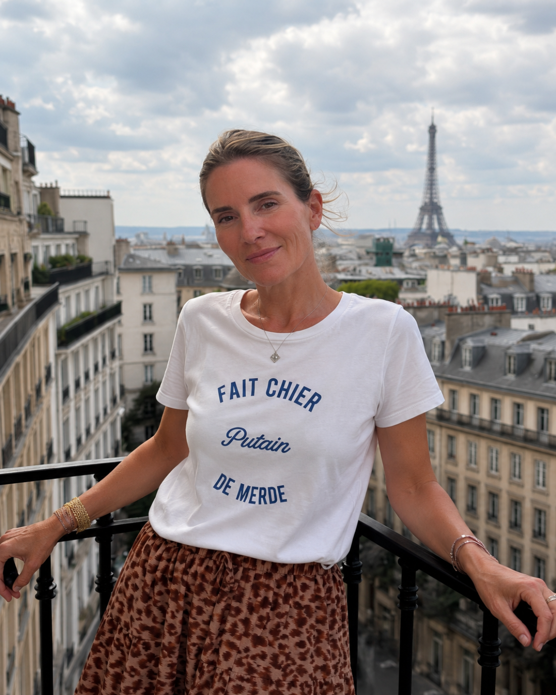 Woman standing on a balcony with the Eiffel Tower in the background, wearing a white t-shirt with blue text.